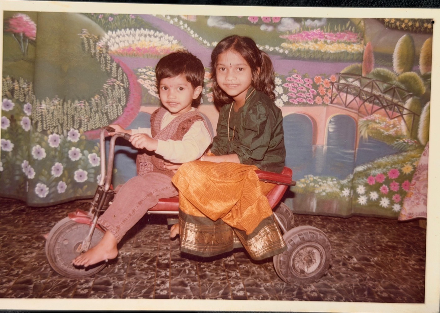 Madhuri and Abhilash as young children on a tricycle at a photo studio